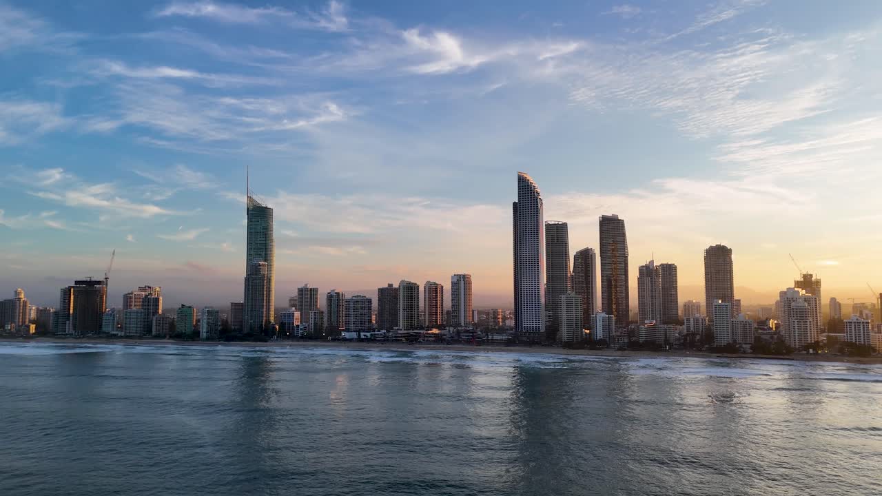 Drone captures Gold Coast skyline transitioning from day to night, highlighting skyscrapers and ocean under vibrant sunset hues