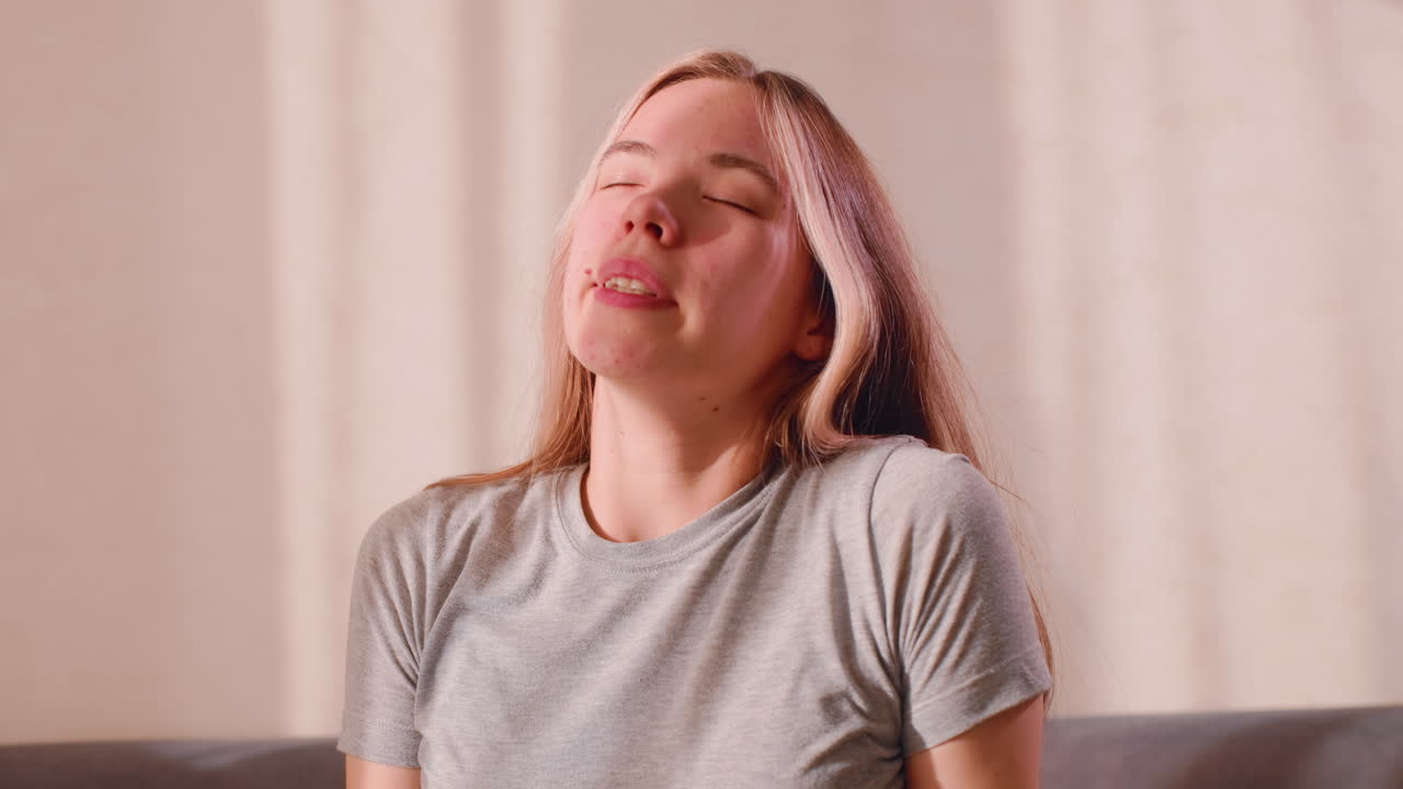 Young woman in grey shirt stretches both arms upward while seated, eyes gently closed, soft sunlight casting warm patterns on wall, expressing calm relief and body awakening