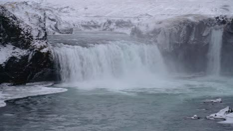 panning shot of godafoss waterfall crashing down during frosty cold day in winter, iceland - icebergs floating in water