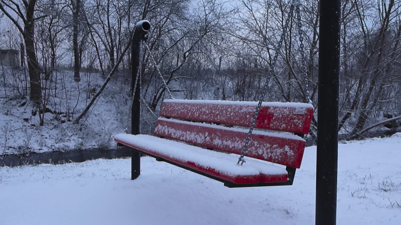 Red colored bench hanging on metal post with chains covered in white fluffy and thick snow in a park in Laagri during winter cloudy day. Dark river water flowing in slow motion in the background.
