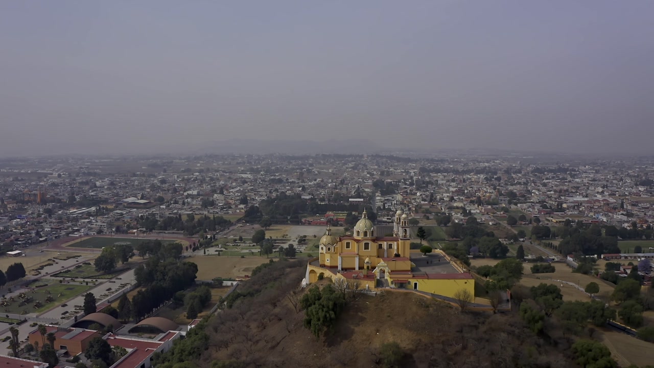 An aerial drone shot captures the iconic Santuario de Nuestra Señora de los Remedios (Church of Our Lady of Remedies) perched atop the Great Pyramid of Cholula in Puebla, Mexico.