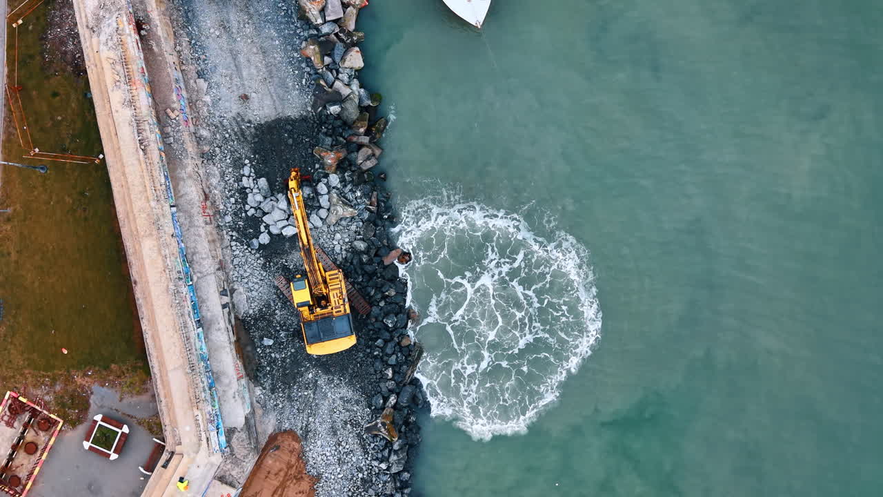 Veduta Aerea dall'Alto di Escavatore al Lavoro sulla Costa. Escavatore giallo che sposta rocce su una costa sassosa con acqua turchese che si infrange.