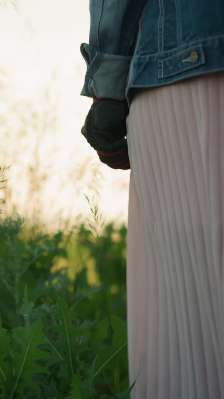 청바지 재과 분홍색 플리티 드론을 입은 여자가 높은 풀을 고 있으며, 그녀의 손은 식물들을 약간 고 있습니다.