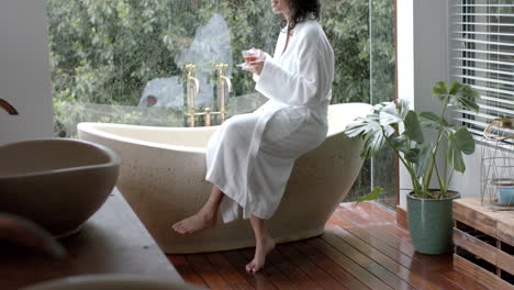 biracial woman wearing bathrobe sitting on bathtub drinking tea in bathroom at home, slow motion