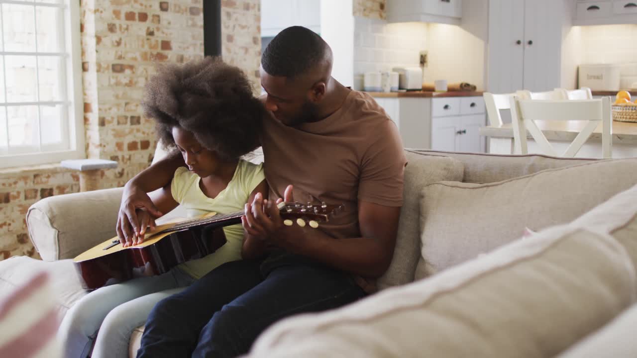 Padre afroamericano e sua figlia seduti sul divano che suonano la chitarra insieme.