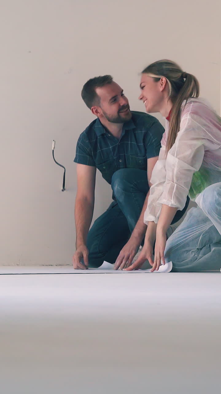lovely young couple unrolls sheet of new wallpaper together smiling and hugging on floor near white wall in renewing room
