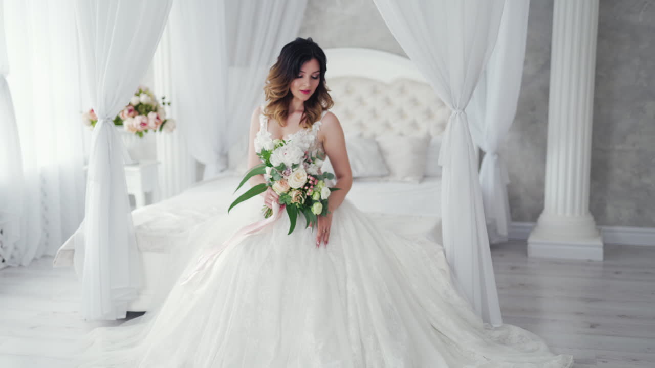Attractive young bride in a sexy wedding dress is sitting on bed. Beautiful bride with flowers. Pretty model in white dress is posing to the female photographer on the bedroom background.