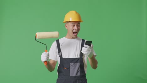 asian man painter wearing safety helmet looking at smartphone, smiling, and saying wow while standing in the green screen background studio