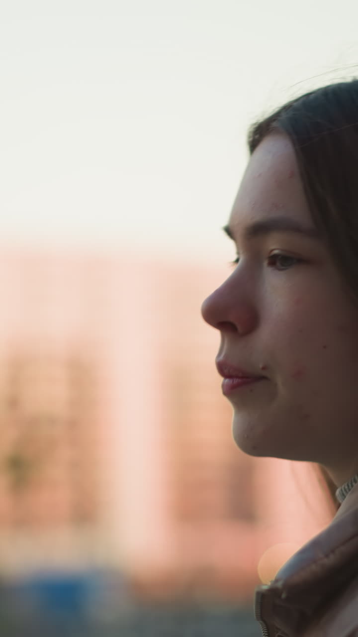 una toma de perfil en primer plano de una joven con una chaqueta de melocotón, caminando por un parque. la toma captura su expresión enfocada y cabello largo, con un fondo urbano borroso
