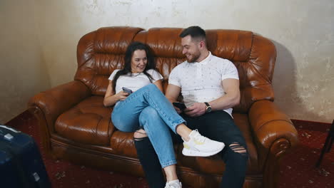 happy young couple on a sofa in hotel. smiling man and woman in casual clothes sitting together with mobile phones and talk on suitcases background.