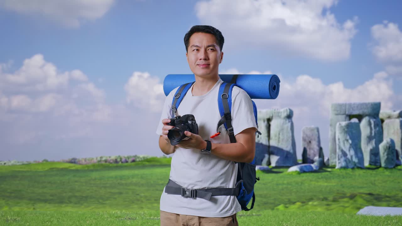 Asian Male Hiker With Mountaineering Backpack Smiling And Holding A Camera In His Hands Then Looking Around While Traveling In Stonehenge