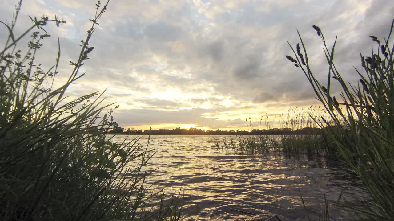A mesmerizing timelapse of the sun descending over a tranquil lake, with light rays breaking through the clouds and tall grasses framing the scene.