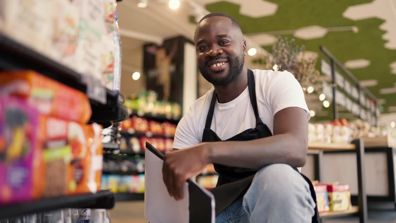 Portrait of a Black-skinned man in a white t-shirt and black apron crouched near the shop window, looks at the camera and smiles