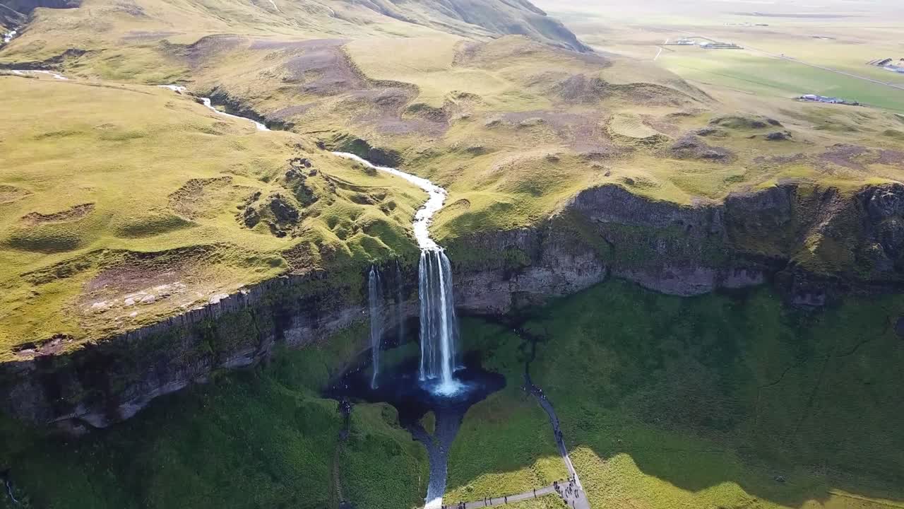 Aerial view of Seljalandsfoss waterfall in Iceland, surrounded by lush green landscapes and cliffs, with a clear river flowing over the edge into a serene pool below