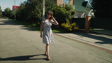 stylish woman in floral dress and sunglasses walks along quiet residential street, adjusting her hair as she passes elegant suburban homes surrounded by greenery and blooming yellow flowers