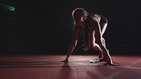 atleta femenina en un fondo oscuro se está preparando para correr el sprint de cross-country desde las almohadillas en la cinta de correr en un fondo oscuro