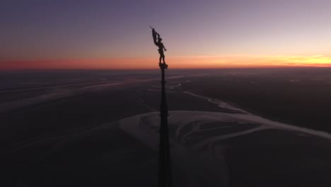 Silhouette-of-statue-on-top-of-Abbey-spire-and-view-of-bay-at-dusk,-Mont-Saint-Michel-in-Normandy,-France