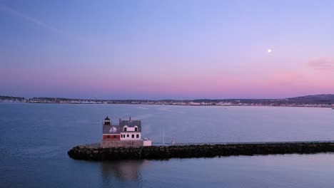 aerial footage flying past the lighthouse at the end of a snow covered rocky breakwater toward rockland maine and a setting full moon