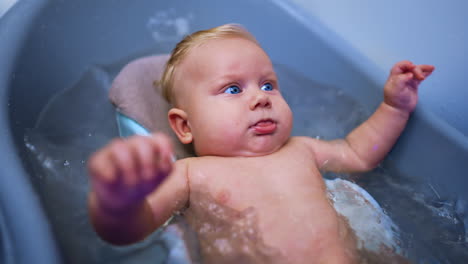cute caucasian infant with blond hair and big blue eyes lies in a bath. beautiful baby bathing close up.
