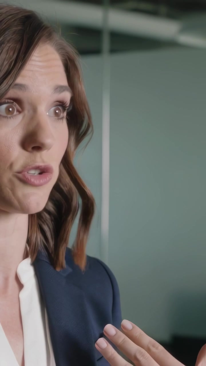 Video verticale: Donna in blazer che parla e risponde in ufficio, gesticolando con due dita verso una parete di vetro.