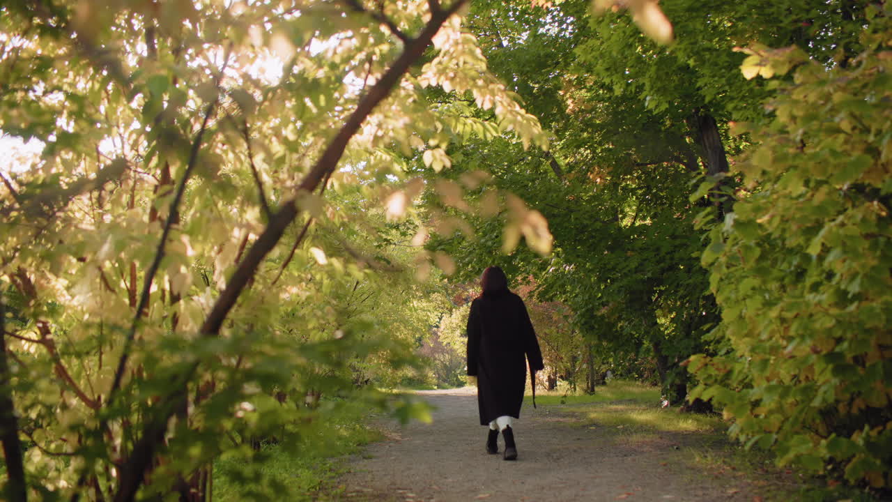 Quiet walker with headphones holds autumn leaf, listens to music and spins around under sunlit arboretum canopy, relaxed stroll along leafy path, golden foliage framing calm outdoor moment