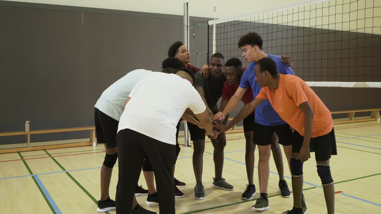 Diverse male teammates huddling at gym net after coach calling rally, chanting, preparing for play