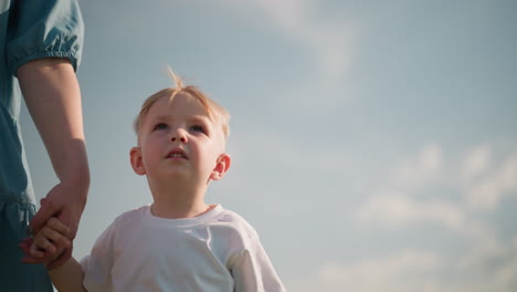 a close-up of a little boy wearing a white shirt, smiling gently while holding his mother s hand as they walk together under a bright sky. the mother is only partially visible