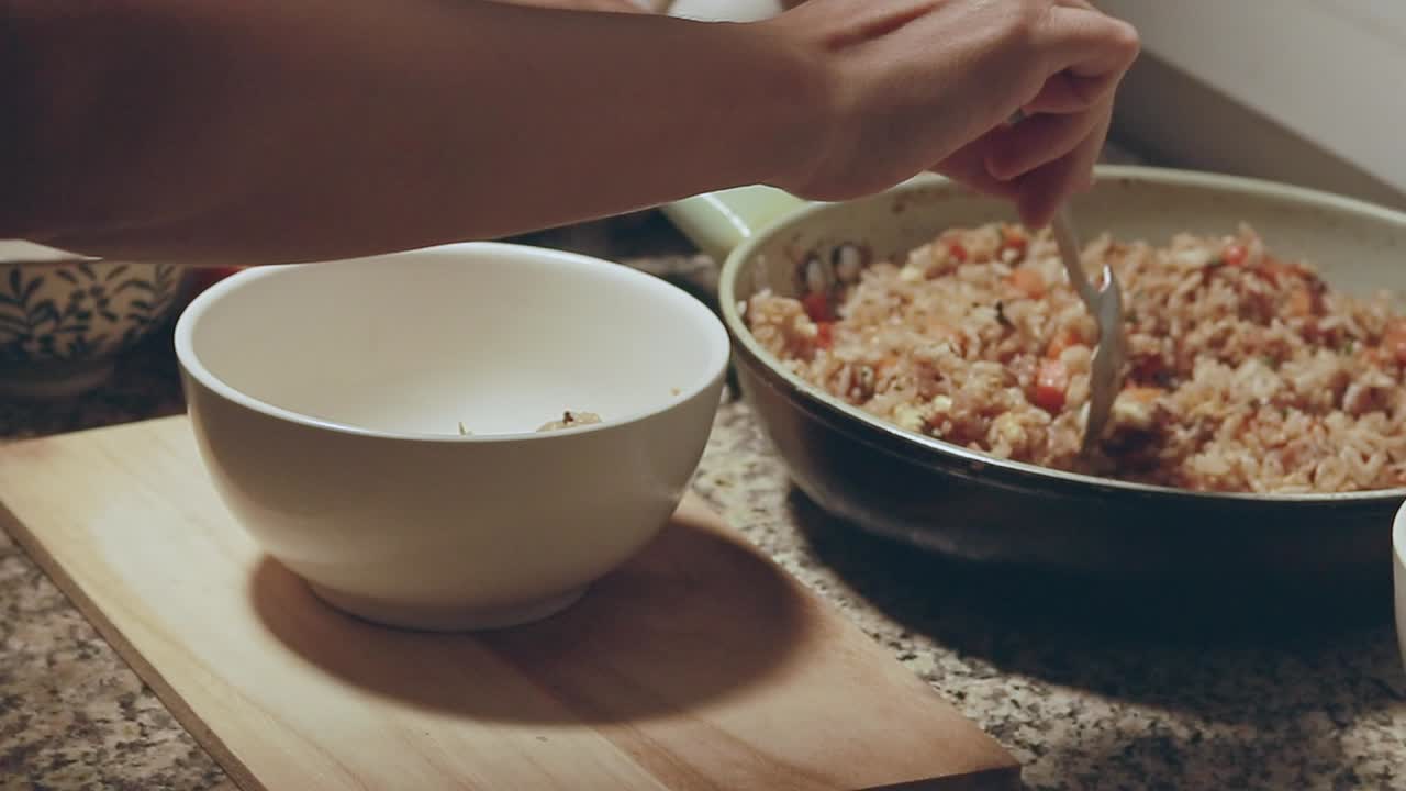 A person serves fried rice from a pan on a kitchen countertop to a white bowl, candid moment of daily domestic home life