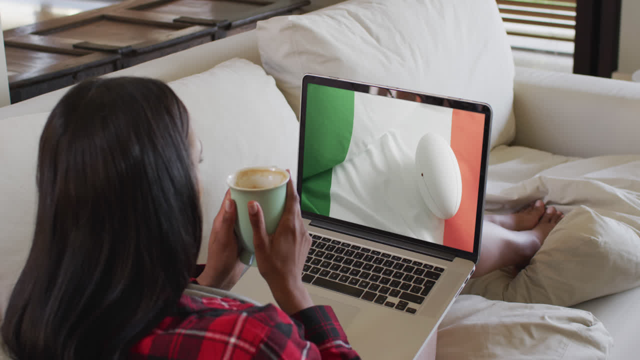 mujer biracial viendo una computadora portátil con una pelota de rugby con la bandera de la costa de marfil en la pantalla