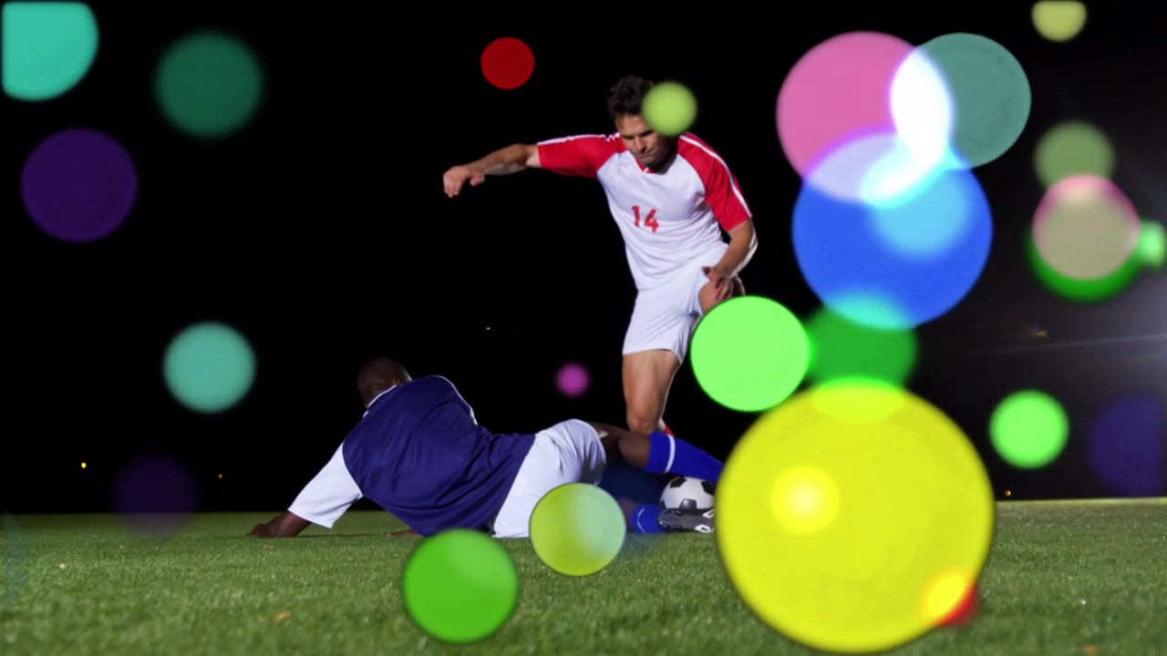 Sudut rendah: pemain nomor 14 berbaju putih-merah menggiring bola, lawan berbaju biru meluncur merebut bola, pemain lain membantu di tengah efek bokeh.