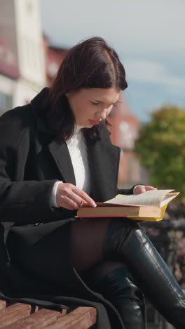 joven sentada al aire libre en un banco leyendo su libro, con un enfoque en las páginas del libro, lleva botas negras y un abrigo mientras una bolsa descansa a su lado, entorno urbano en el fondo