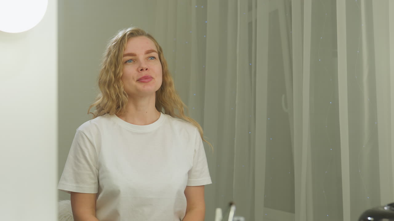 Blond woman in white shirt sitting in front of illuminated mirror smiling calmly while beauty tools and products are arranged on salon table with curtain backdrop and soft lighting ambiance