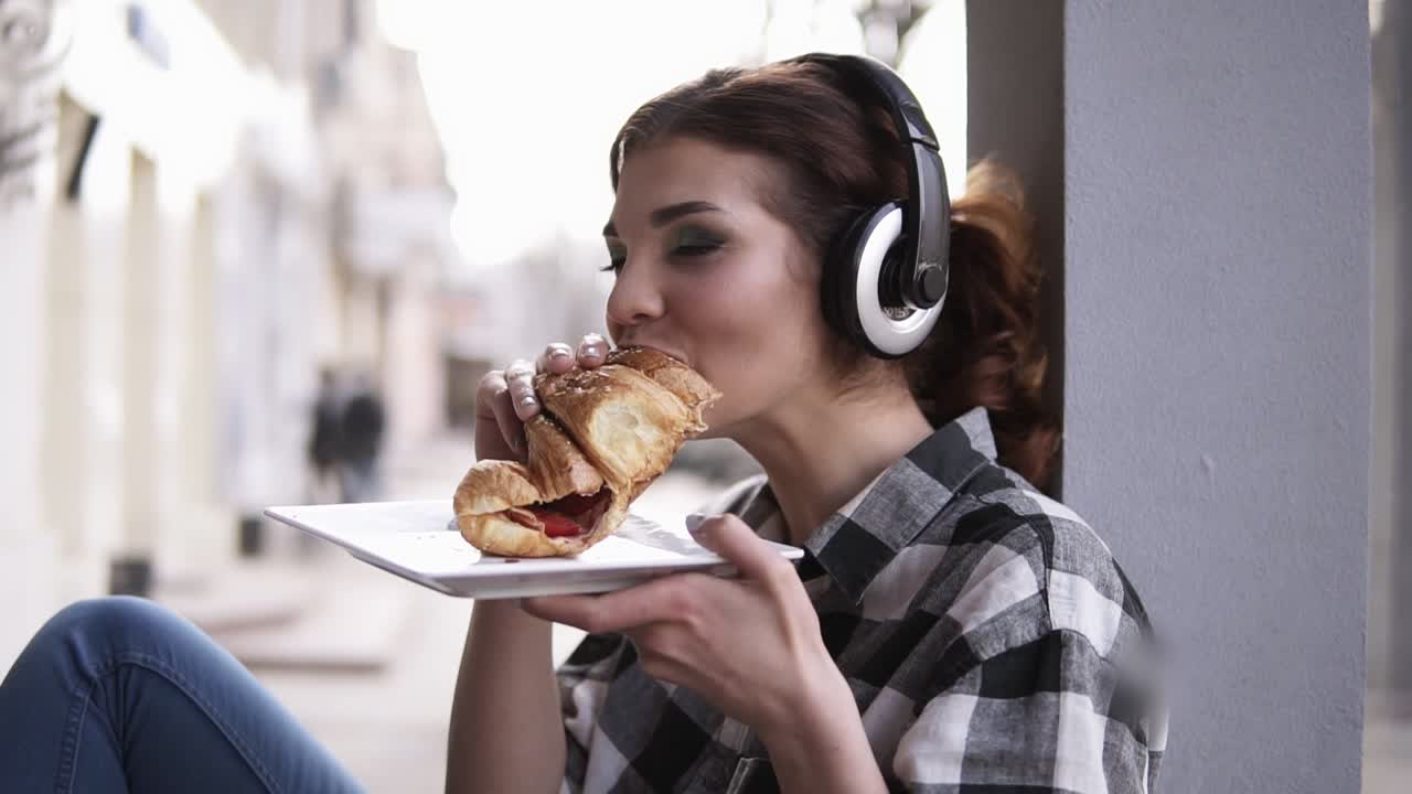 hermosa, joven sentada junto a la ventana con grandes auriculares. escuchando música y comiendo un croissant. sosteniendo un plato en sus brazos. feliz, sonriente. cámara lenta