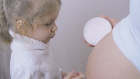 blond girl applies lotion on pregnant mother belly from jar