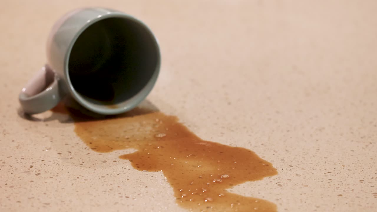 A hand uses a spray bottle to clean a coffee spill from a tipped mug on a table