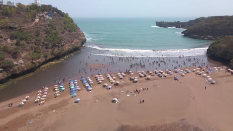 Aerial-view-of-sandy-beach-is-busy-with-people-playing-in-the-sea-waves-and-colorful-umbrellas-set-up-on-the-sand-for-shade---Baron-beach,-Indonesia