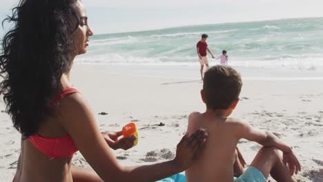 vista de atrás de la madre hispana aplicando crema solar en el hijo de vuelta en la playa