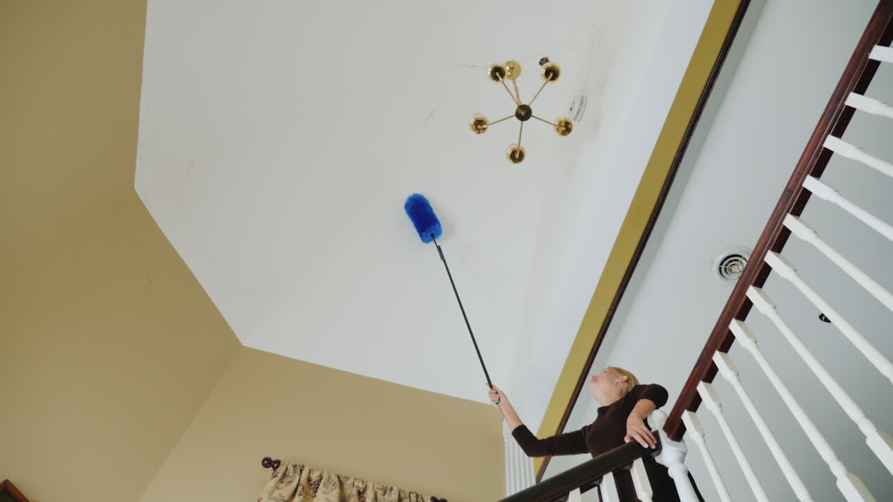 una mujer limpia las telarañas y los escombros en el techo cerca de la limpieza de la casa grande de la lámpara de araña