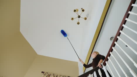 a woman cleans the cobwebs and debris on the ceiling near the chandelier big house cleaning