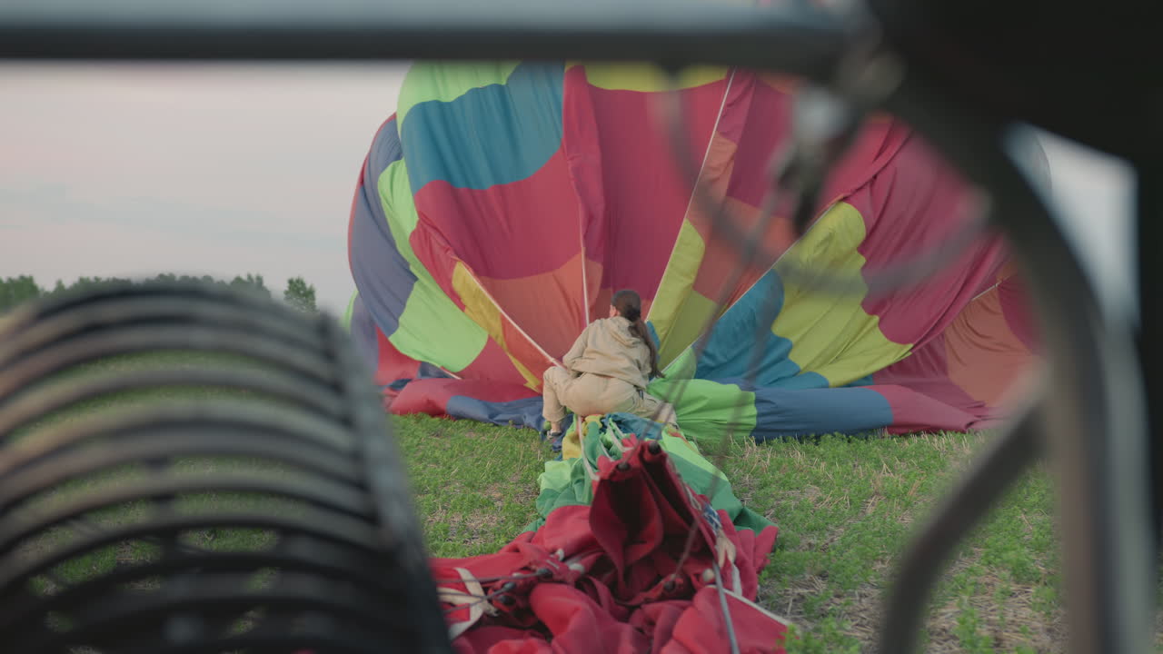 close up of metal burner with back view of woman in beige suit crouching on green field folding vibrant hot air balloon fabric after landing during peaceful evening under soft pastel sky
