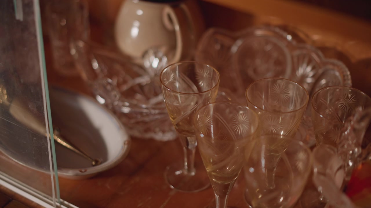 Små likørglas i et træskab, kurator arrangerer krystalglas med stilk ved siden af en tekande og skåle af porcelæn, blødt ravfarvet lys fremhæver skrøbelige, udsmykkede detaljer og reflekterende glans.