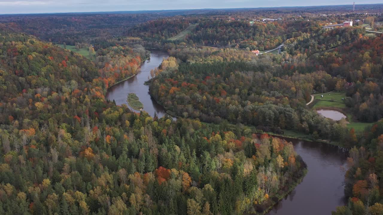 A drone rises above Sigulda’s lush forest, unveiling the winding Gauja River as it snakes through a tapestry of vibrant trees, with the scenic valley and stunning natural landscape unfolding below