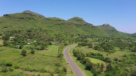 A-drone-shot-of-green-farmlands-and-a-highway-cutting-through,-a-range-of-hills-on-one-side-and-a-motorcycle-riding-on-the-highway