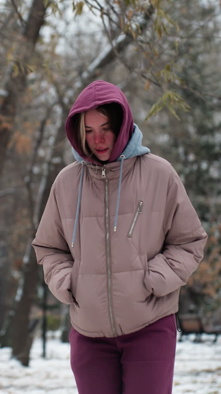 따뜻한 후디와 코트를 입은 여자가 겨울 스노우 파크에서 장난스럽게 점프합니다. 배경에 눈으로 인 나무는 평화롭고 경치 좋은 분위기를 만니다.