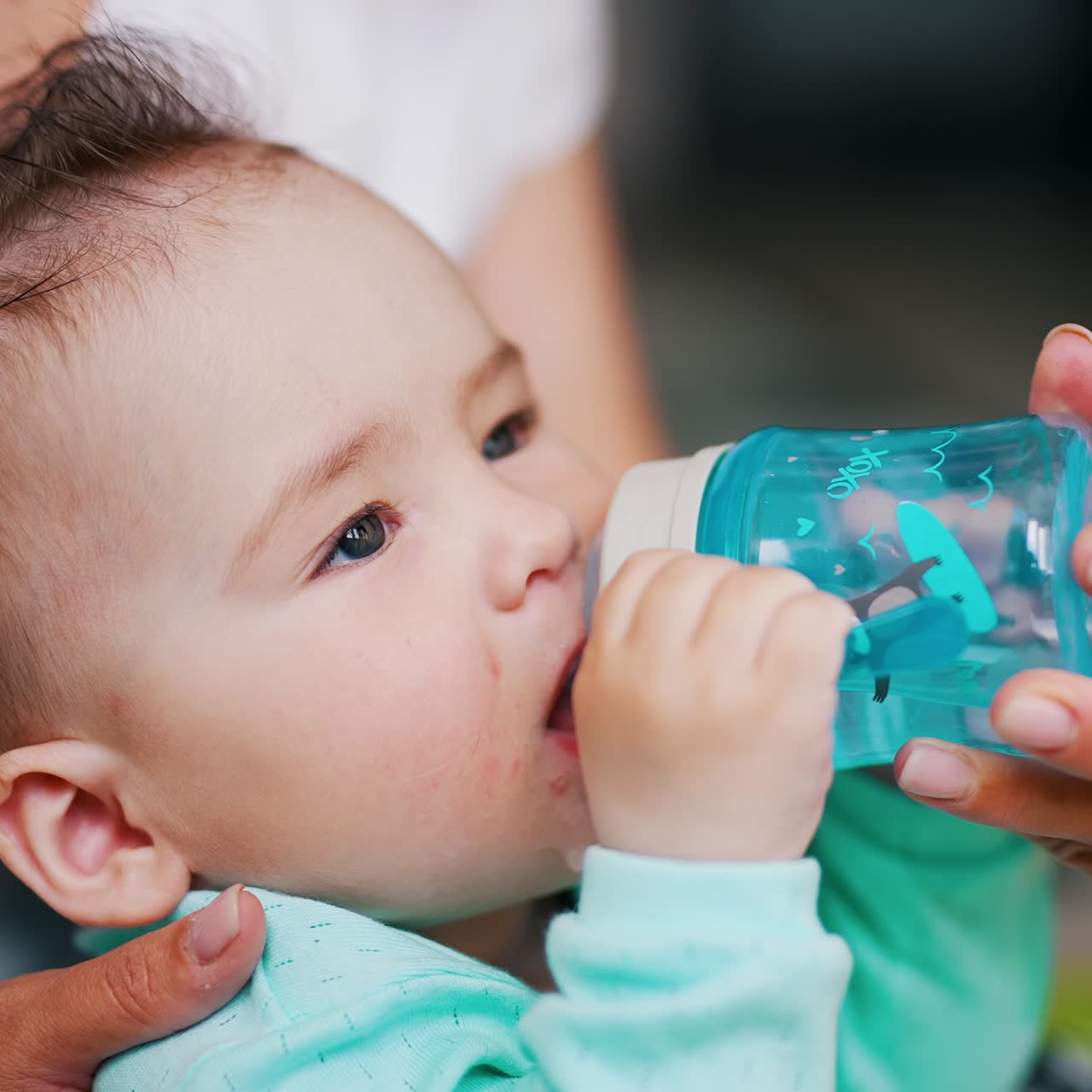 Lovely toddler with stylish hair drinking water from a bottle. Mom's hand is holding a bottle for her kid. Close up