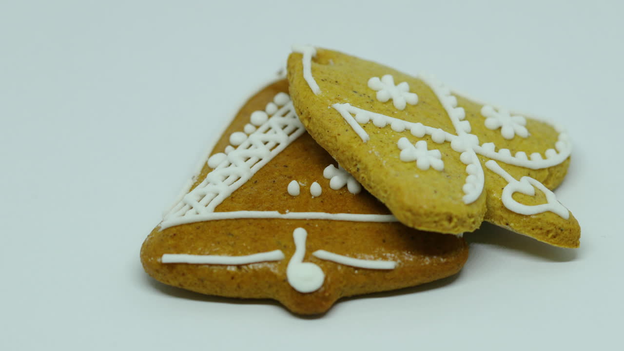 Carrellata laterale su due pan di zenzero con decorazione bianca a forma di campana e cuore su sfondo bianco.