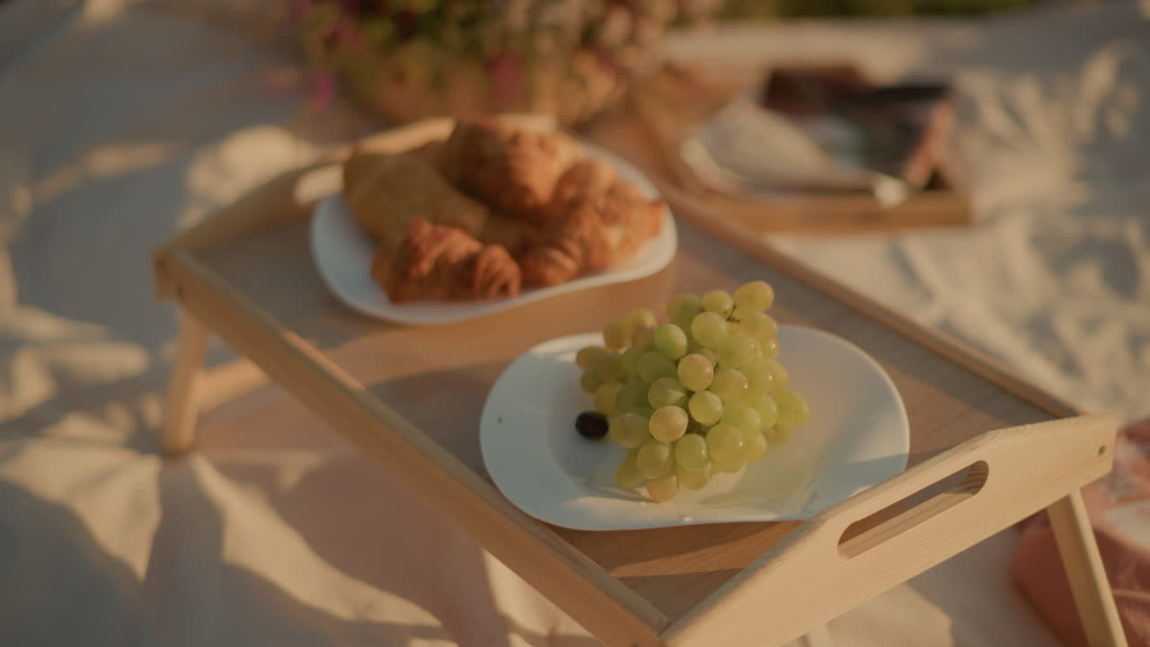 primer plano de la configuración de picnic en una mesa de madera con platos de uvas verdes frescas, croissants y bocadillos bajo la luz solar natural, los platos están artísticamente dispuestos creando un aire libre relajado