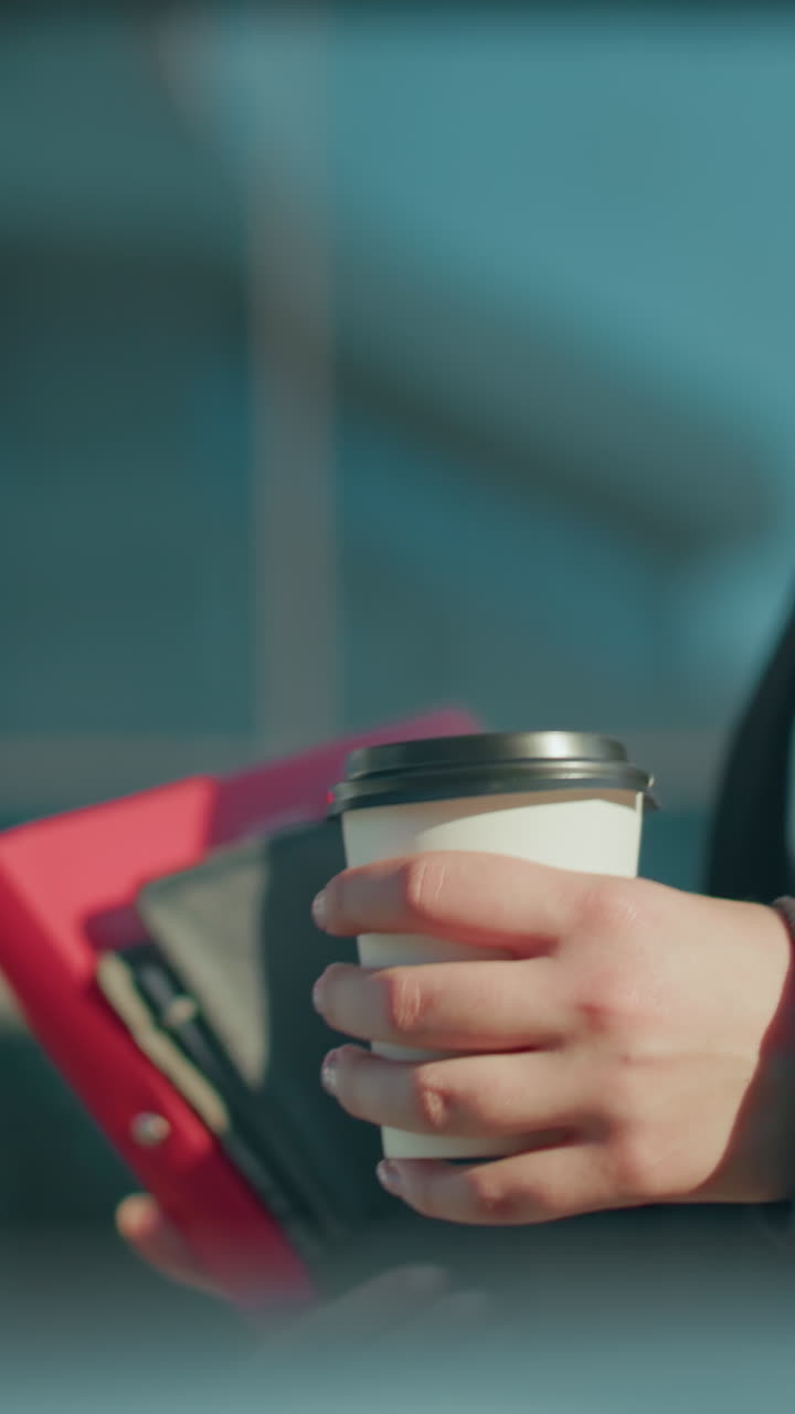 Close up side view of woman in formal wear walking outdoors while holding file folder and coffee cup with bright sunlight creating ambient reflections on metal railing and modern glass building