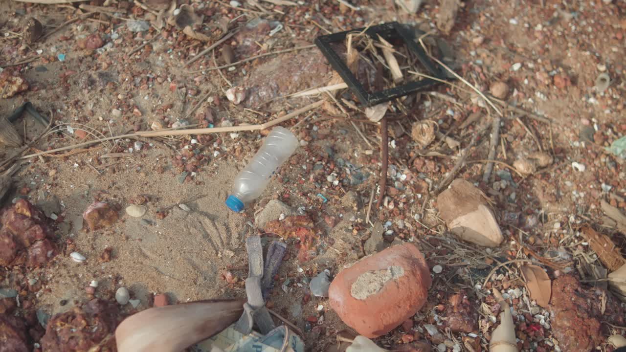 Rotating ground-level shot showing plastic bottles, trash, and rubble scattered on sandy ground near the shoreline