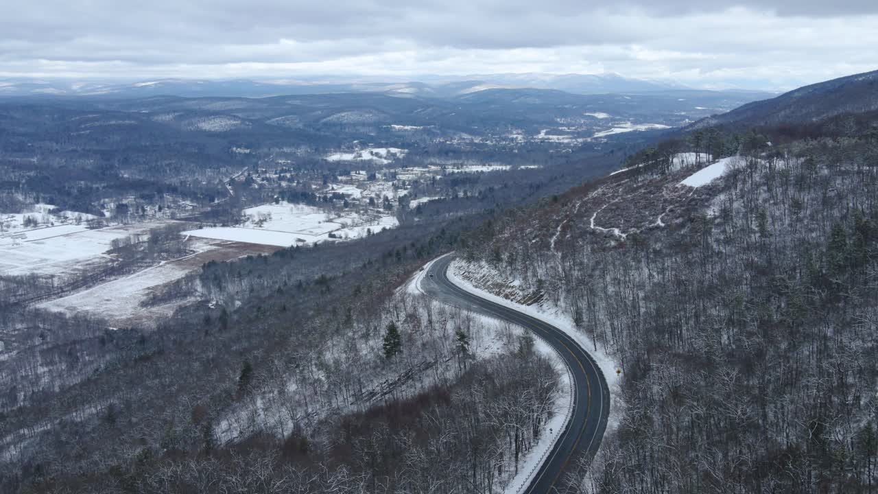 Riprese aeree con drone di una strada panoramica tra montagne innevate, con una vasta montagna innevata sottostante e cime innevate in lontananza.